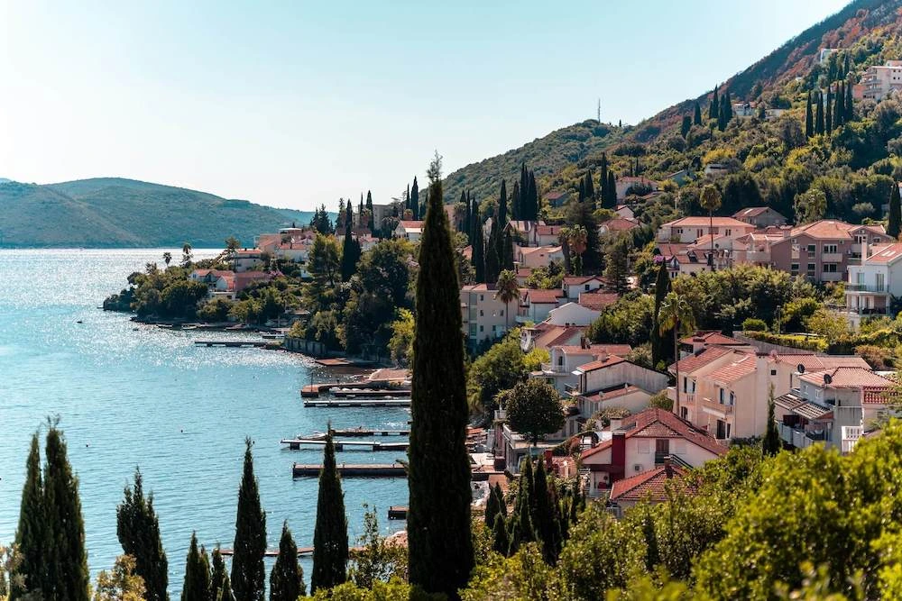 Image of a hillside sloping down to the Adriatic sea in Montengro, with houses and trees clinging to the hill, clear blue skies and shimmering water.
